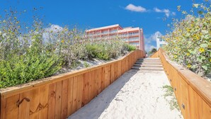 Une promenade en bois mène à un bâtiment doté de nombreux balcons, qui est lui-même entouré d’un terrain sablonneux et de végétation.