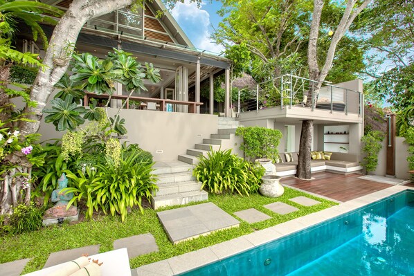 Una casa moderna con piscina, rodeada de exuberante vegetación y un espacio exterior cubierto.
