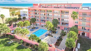 A pink hotel with multiple balconies, a swimming pool, and palm trees. - Beachfront Boutique Resort | Beachfront Bliss | Casa Condo 103 (Port Aransas)