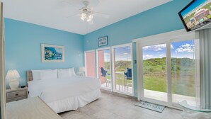 A bedroom with a bed, a nightstand, a lamp, a ceiling fan, a TV, and a view of the outdoors through large sliding glass doors. - Beachfront Boutique Resort | Beachfront Bliss | Casa Condo 103 (Port Aransas)