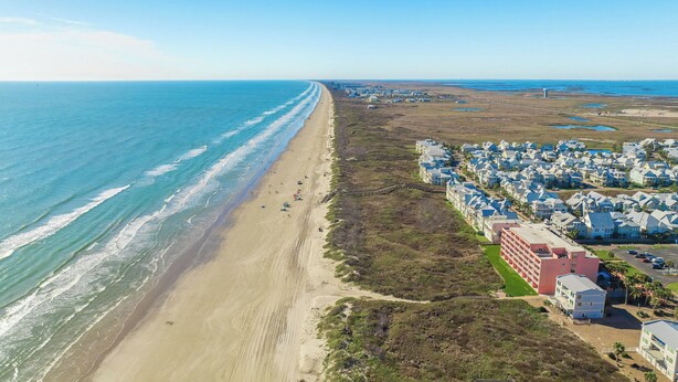 An aerial view of a coastal area with a long sandy beach, a row of residential buildings, and a clear blue sky. - Beachfront Boutique Resort | Beachfront Bliss | Casa Condo 103 (Port Aransas)