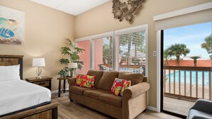 A bedroom with a bed, a sofa, a nightstand, a lamp, a plant, and a view of a pool through a sliding glass door.