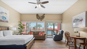 A spacious room with a bed, a sofa, a dining table with chairs, a ceiling fan, and a large window with blinds.