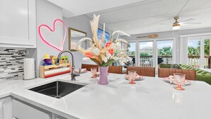 Una cocina moderna con encimera blanca, un fregadero y un florero. Hay una zona de comedor con una mesa preparada para dos personas, y vistas al exterior a través de amplias ventanas.