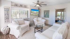 A spacious living room with white furniture, a ceiling fan, and a large window with a view of palm trees. - Beachfront Boutique Resort |Serene Escape| Casa Condo 210 (Port Aransas)
