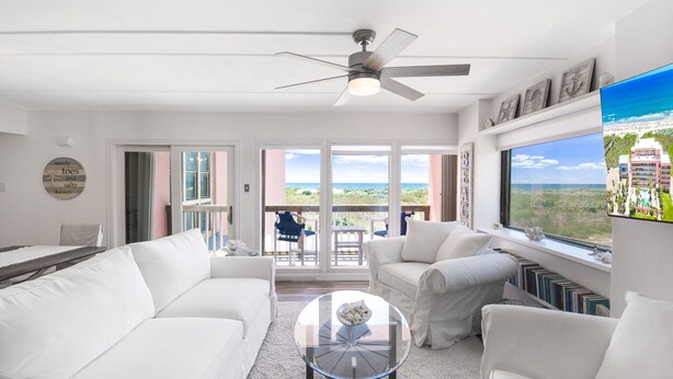 A spacious living room with white furniture, a ceiling fan, a glass coffee table, and a view of the outdoors through large windows. - Beachfront Boutique Resort |Serene Escape| Casa Condo 210 (Port Aransas)