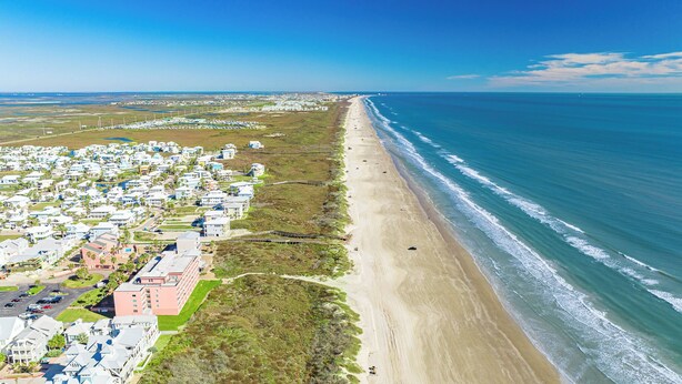 Vista aérea de una zona costera con una playa extensa, un barrio residencial con casas y un cielo azul despejado.