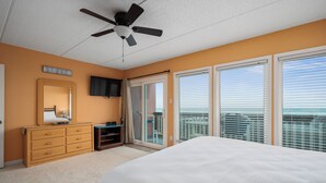Un dormitorio con una cama grande, ventilador de techo, cómoda, televisor y puerta corrediza de vidrio que da a un balcón.