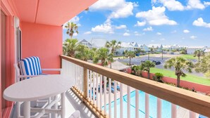 Un balcón con una mesa blanca y una silla a rayas, con vistas a una zona de piscina con palmeras y un aparcamiento.