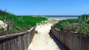 Ein sandiger Pfad führt zu einem Strand, der beidseitig von Holzzäunen gesäumt ist.