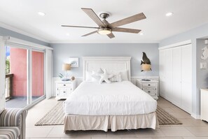A bedroom with a large bed, a ceiling fan, a nightstand with a lamp, and a view of the outside. - Beachfront Boutique Resort | Sunkissed Retreat | Casa Condo 307 (Port Aransas)