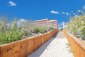 Une promenade en bois menant à un bâtiment doté de balcons, entouré d’un terrain sablonneux et de végétation.