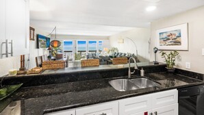 Una cocina moderna con encimera de granito negro, pileta de acero inoxidable y alacenas blancas. Hay un comedor con vista al océano a través de amplias ventanas.