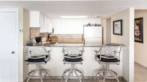A kitchen with white cabinets, a white refrigerator, a coffee maker, and three bar stools. - Beachfront Boutique Resort | Sunset Serentity | Casa Condo 402 (Port Aransas)