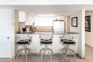A kitchen with white cabinets, a white refrigerator, a coffee maker, and three bar stools.