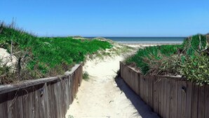 A sandy pathway flanked by wooden fences leading to a beach. - Beachfront Boutique Resort | Sunset Serentity | Casa Condo 402 (Port Aransas)