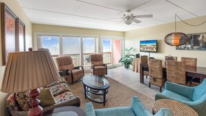 A spacious living room with a ceiling fan, a television, and a view of the outdoors through large windows. - Beachfront Boutique Resort | Sunset Serentity | Casa Condo 402 (Port Aransas)