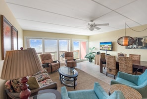 A spacious living room with a ceiling fan, a television, and a view of the outdoors through large windows.