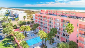 A pink hotel with a pool, surrounded by palm trees and a beach view.