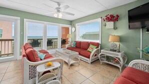 A sunlit room with a ceiling fan, wicker furniture, and a view of the ocean through large windows.