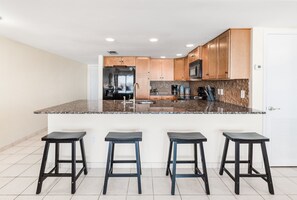 A modern kitchen with a granite countertop, stainless steel appliances, wooden cabinets, and three black stools. - Beachfront Boutique Resort | Oceanside Paradise | Casa Condo 501 (Port Aransas)