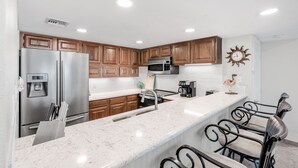 A modern kitchen with wooden cabinets, stainless steel appliances, a marble countertop, and a bar area with metal chairs.