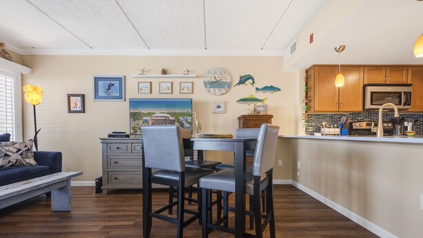 A dining area with a table and chairs, a kitchen with wooden cabinets and a microwave, and a living room with a sofa and a lamp. - Beachfront Boutique Resort | Pelican's Perch | Casa Condo 506 (Port Aransas)