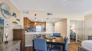 A compact kitchen with modern appliances, a dining area with blue chairs, and a bedroom with a bed and nightstand. - Beachfront Boutique Resort | Pelican's Perch | Casa Condo 506 (Port Aransas)