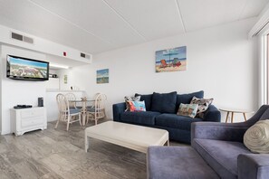 A modern living room with a blue sofa, a white coffee table, and a dining area with wicker chairs.