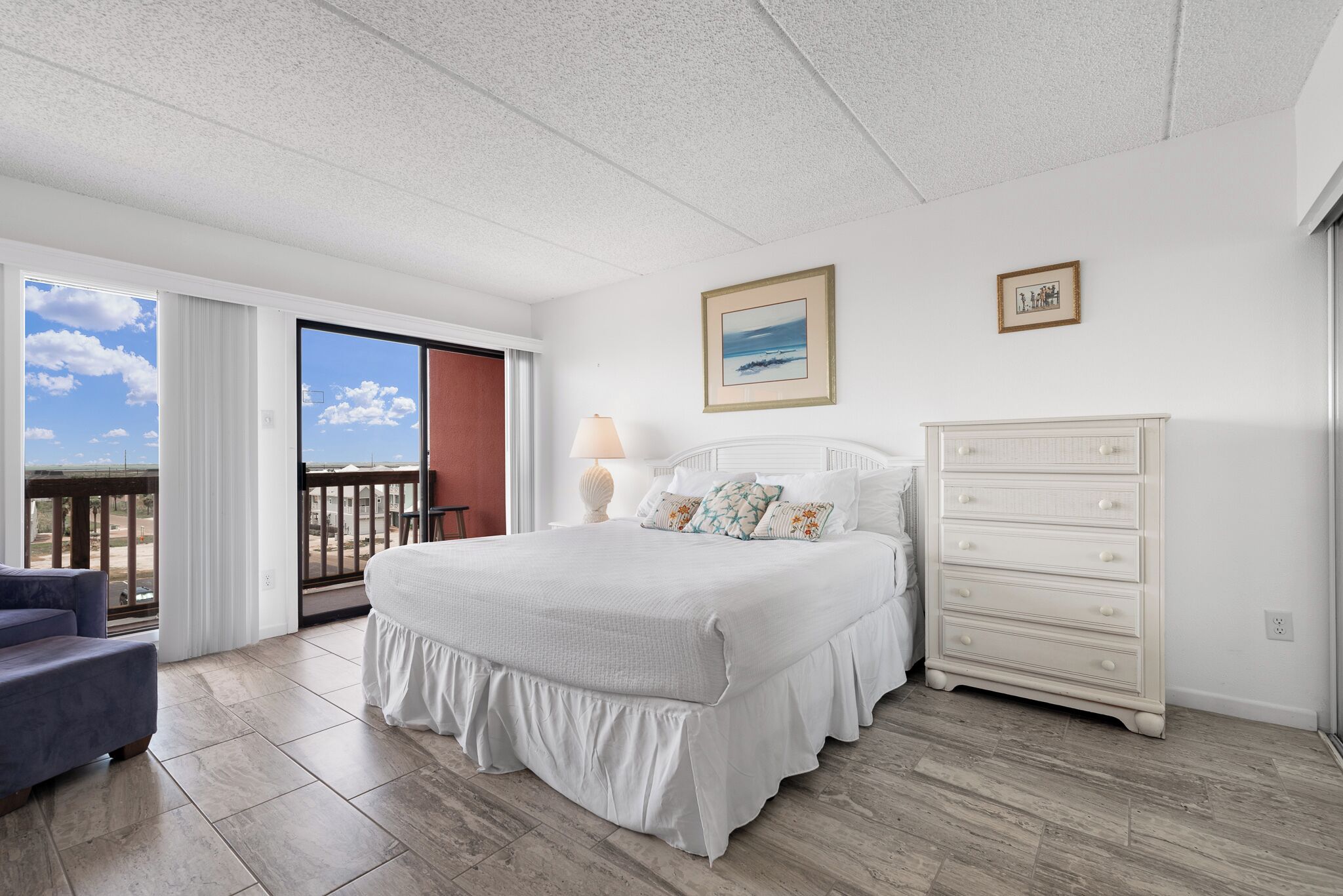 A bedroom with a large bed, a dresser, a chair, and a balcony with a view.