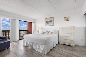 A bedroom with a large bed, a dresser, a chair, and a balcony with a view.