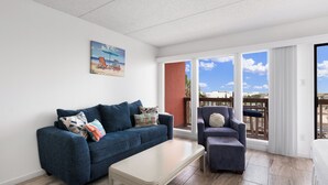 Ein Wohnzimmer mit einem blauen Sofa, einem weißen Couchtisch und einem blauen Sessel. An der Wand hängt ein Gemälde und durch große Fenster hat man einen Blick nach draußen.