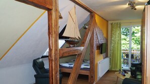 A loft-style room with a wooden beam structure, a sailboat model, a green sofa, and a view of the outdoors through a glass door. - Segelwohnung - Meerzauber. Gutshaus Reidervitz - Großer Lindengarten (Altenkirchen)