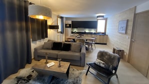 Ein modernes Wohnzimmer mit einem grauen Sofa, einem Couchtisch und einem kuscheligen Sessel. Es gibt einen Essbereich mit Tisch und Stühlen, und im Hintergrund eine Küche.