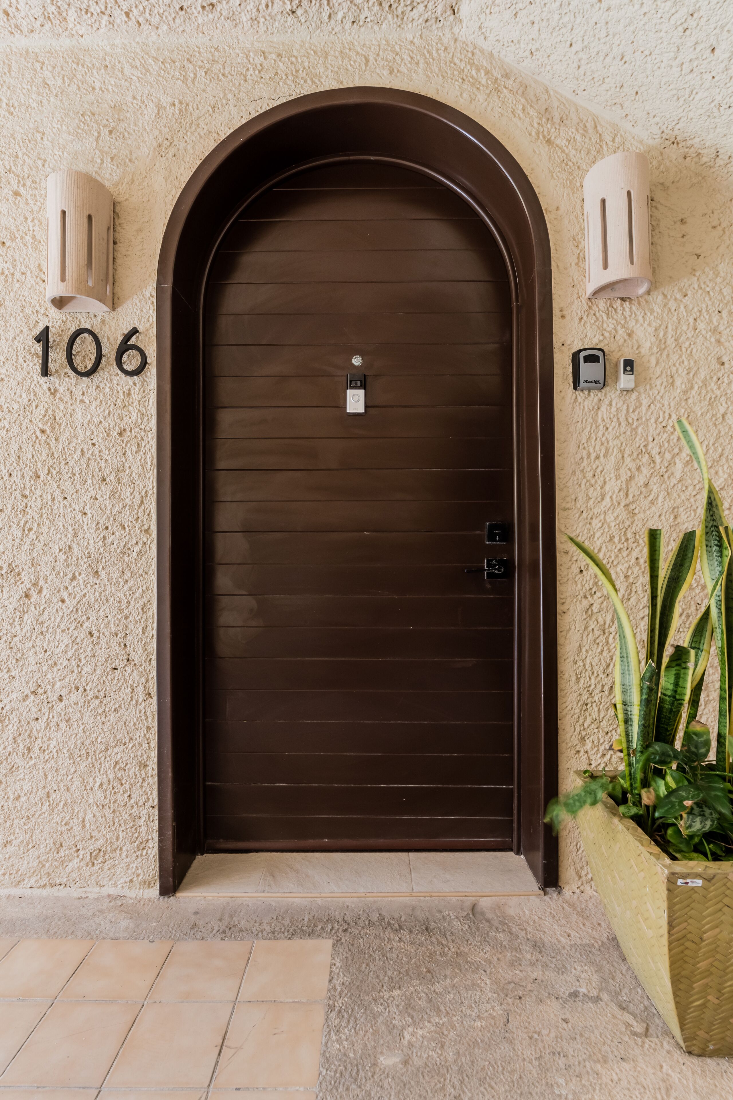 Une porte marron avec le numéro « 106 » au-dessus, une petite boîte aux lettres et un jardon rempli de plantes vertes.