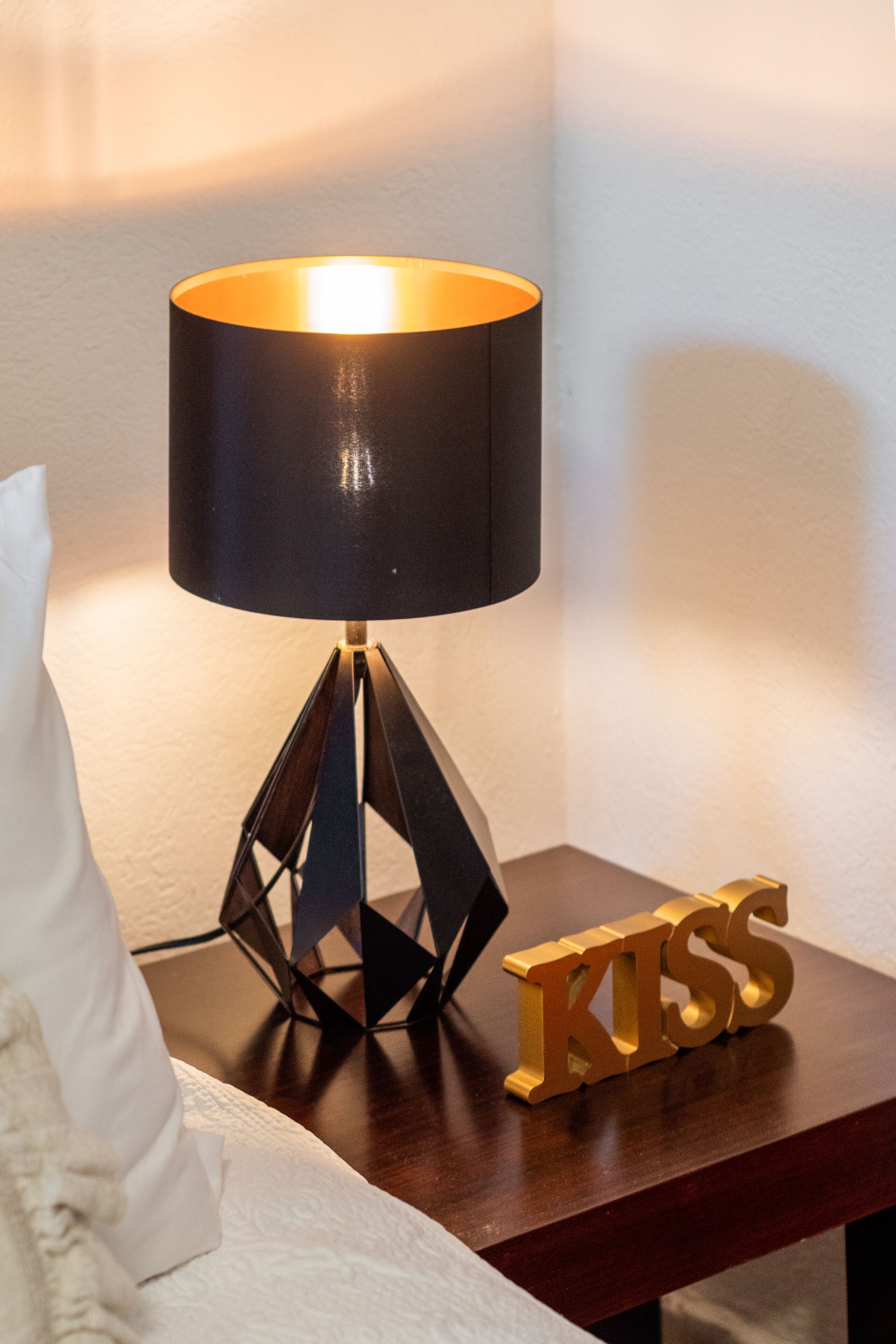 Een nachtkastje met een zwarte lamp, een wit kussen en een gouden bord met de tekst 'KISS'.
