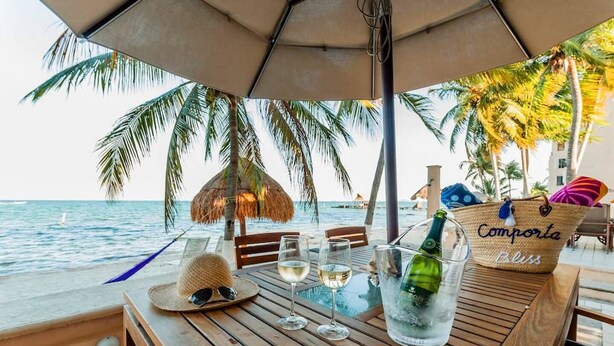 A wooden table with a bottle of champagne, glasses, and a straw hat on a beach setting with a view of the ocean and palm trees. - Depa Bliss Cancún • Beachfront · King Beds · Pool · Luxury Stay • Best Host (Cancún)
