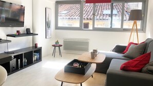 Ein modernes Wohnzimmer mit einem grauen Sofa, einem Holztisch, einem Fernseher an der Wand und einem Fenster mit Blick auf ein Ziegeldach.