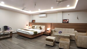 A modern hotel room with a large bed, a sofa, a coffee table, and a desk with a chair. There are ceiling fans, recessed lighting, and an air conditioning unit. - Hotel Atul Residency (Satna)