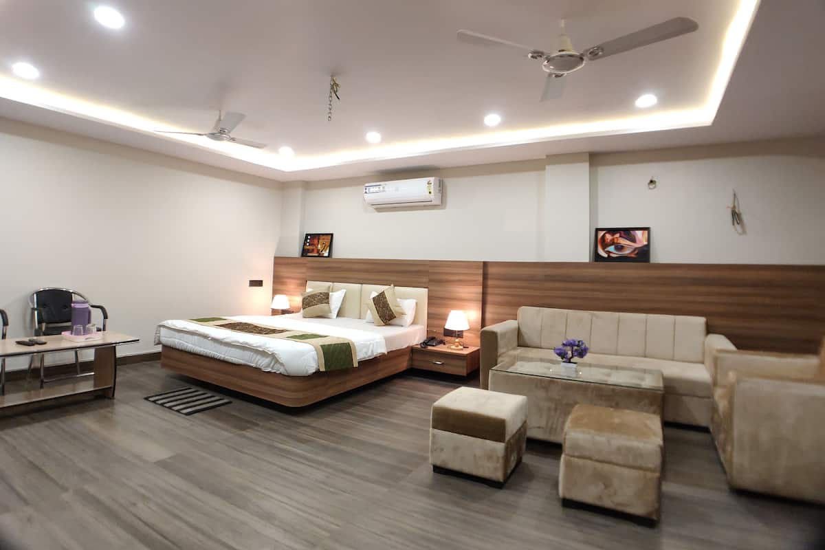 A modern hotel room with a large bed, a sofa, a coffee table, and a desk with a chair. There are ceiling fans, recessed lighting, and an air conditioning unit.