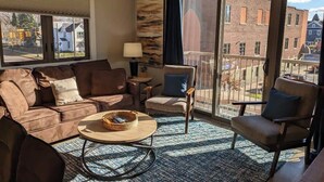 Ein Wohnzimmer mit einer Couch, Sesseln, einem Couchtisch und Blick auf ein Backsteingebäude durch große Fenster.