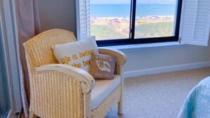 Ein Korbstuhl mit zwei Kissen, eines mit Schrift und das andere mit einer Illustration eines Strandhauses, in einem Zimmer mit einem großen Fenster mit Blick auf einen Strand.