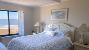 Ein Schlafzimmer mit Bett, Nachttisch, Lampe und Fenster mit Blick auf den Strand.