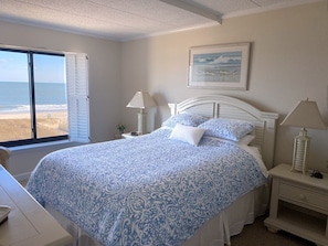 Quarto com cama, mesa de cabeceira, abajur e janela com vista para a praia.