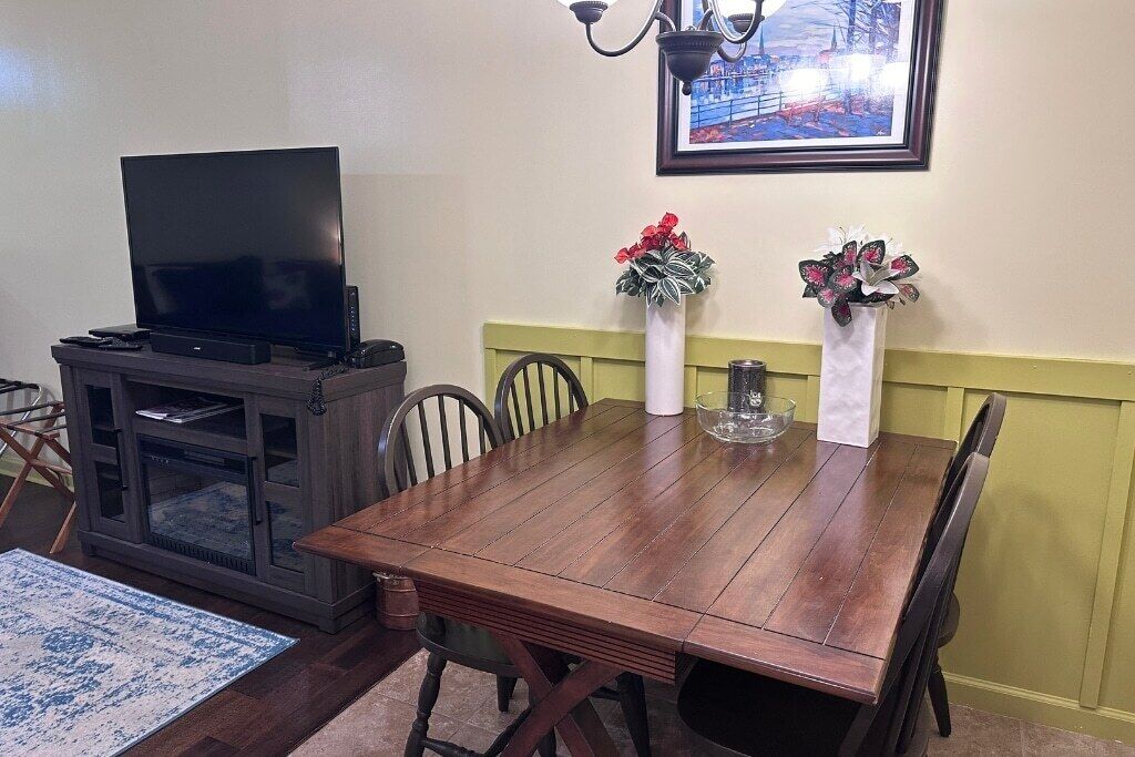 A dining area with a wooden table, chairs, a television on a cabinet, and two vases with flowers.