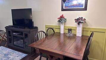 A dining area with a wooden table, chairs, a television on a cabinet, and two vases with flowers.