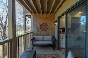 Un balcon avec un canapé en osier, une table en bois et une cuisine visible à travers une porte vitrée.