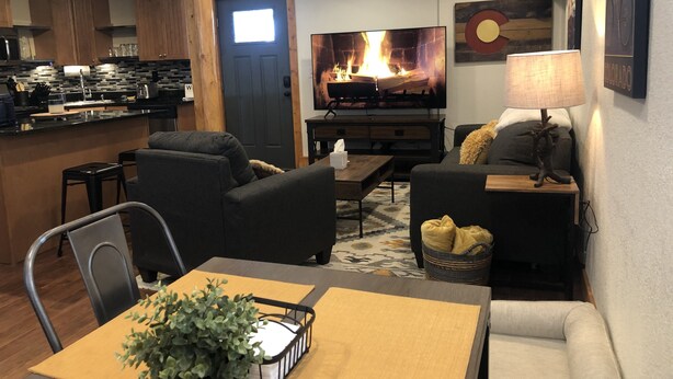 A modern living room with a fireplace, a dining area with a table and chairs, and a kitchen area with wooden cabinets and a bar.