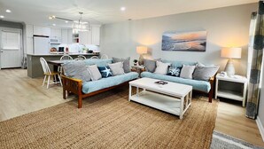 A modern living room with a sofa, coffee table, and lamps. A kitchen is visible in the background. - Vast Views | Remodeled Townhome, Pool, Pier Access, Lighthouse Views + Pets (Chincoteague Island)