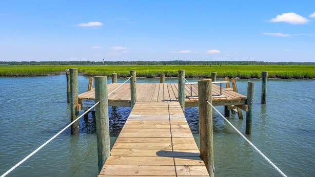 A wooden pier extending into a body of water with a grassy landscape in the background. - Vast Views | Remodeled Townhome, Pool, Pier Access, Lighthouse Views + Pets (Chincoteague Island)